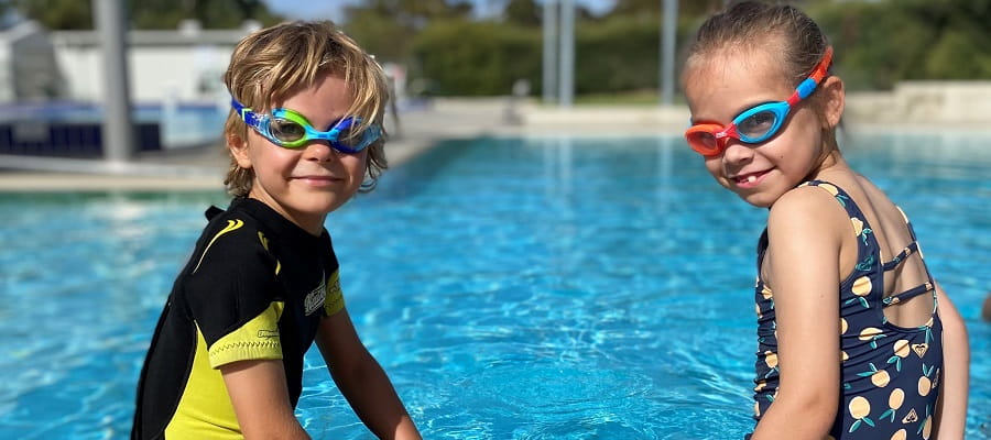 two preschoolers sitting on the edge of a pool smiling at camera