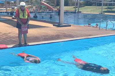 An instructor by the pool with two students floating on their back with red kickboards