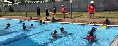 Aboriginal children in the water with others along the edge holding pools noodles to rescue them, instructors looking on
