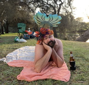 A man dressed as a mermaid laying on a blanket by the river