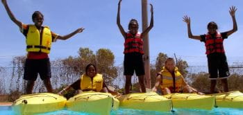 Three indigenous kids playfully balance on kayaks on Bidyadanga pool