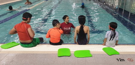 Four children sitting along the edge of a pool facing their instructor in the water