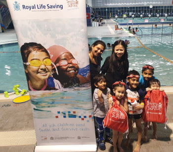 Two mums with their children in bathers with Swim and Survive bags, standing beside a Swim and Survive Access and Equity sign by the pool at Cannington