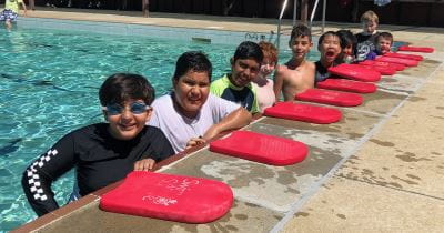 A group of multicultural boys standing in the pool along the edge with red kickboards