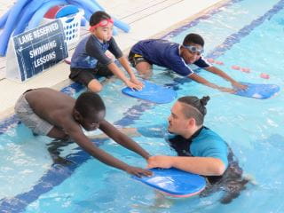 Three boys getting into the pool with kickboards as their instructor assists