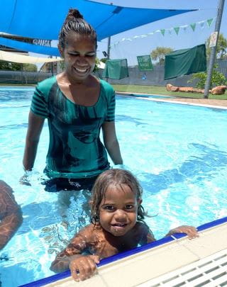 An aboriginal mum standing behind her toddler while he holds the edge of the pool and smiles