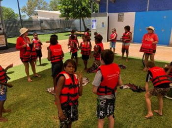 A group of Aboriginal children wearing lifejackets on the grass by the pool at Fitzroy Crossing, with their teaching assisting them