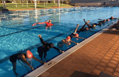 Women lined up along the edge of the pool practising their freestyle breathing while their swim instructor stands behind them in the water