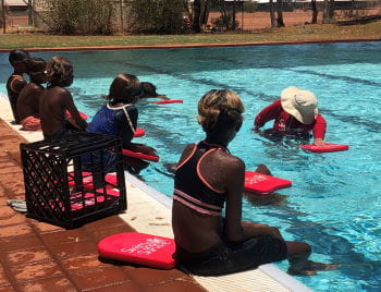 Aboriginal children sitting along the edge of a pool watching their instructor demonstrate freestyle in the water