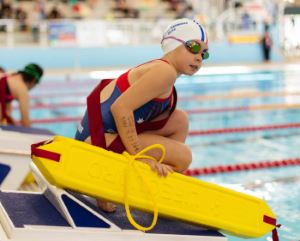 A girl wearing goggles and a swim cap, holding a rescue tube while kneeling on the starting blocks by the pool