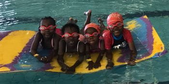 Four children wearing goggles and swim caps laying on a foam mat and kicking around the pool