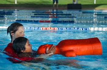 A child holding a pool lifesaving mannequin int he pool with an instructor assisting