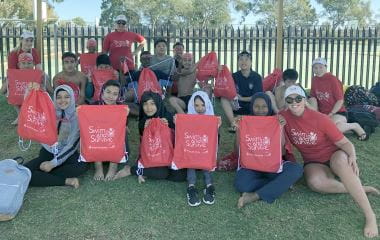 A group of multicultural students sitting on the grass with their swim instructors, holding up red Swim and Survive packs