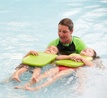 A swim instructor helping two children to float on their back with green kickboards