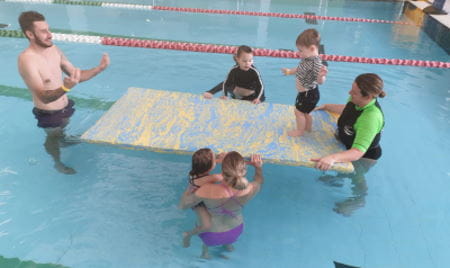 Parents and children in the water with their babies and a swim instructor, with a child walking across a foam mat