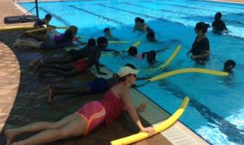 children and teachers from Muludja School laying on the edge of the pool using yellow pool noodles to rescue others in the water