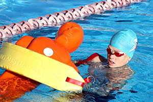A child wearing a swim cap pulling an orange manikin with a rescue tube