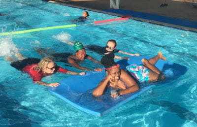 Multicultural women swimming with their instructor in the pool at South Hedland, pushing another woman who is laying on a mat