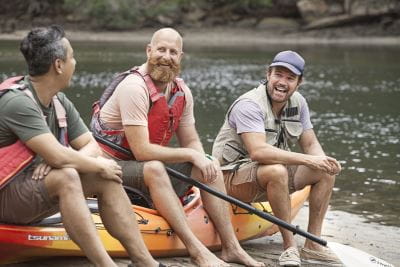 Three guys sitting on a kayak wearing lifejackets and laughing by the river