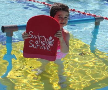 A young girl in the pool holding a Swim and Survive kickboard