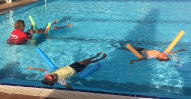 three children floating on their backs using pool noodles with swim instructor assisting