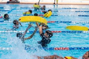 Children in each pool lane putting lifeguard rescue tubes around another child to tow to the end of the pool. 
