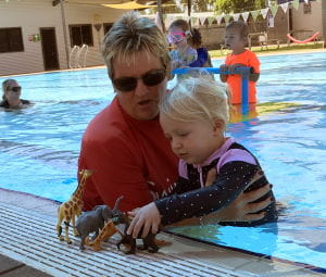 Instructor Adele Caporn with a toddler girl playing with dinosaur toys at the edge of the pool