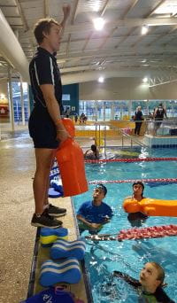 A lifeguard trainer standing on the edge of a pool demonstrating while children gather in the water below