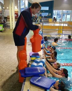 A lifeguard trainer holding a tow manikin by the pool with kids in the water