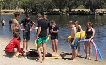 An instructor on the sand by the river with children watching as she demonstrates how to use a spine board to rescue a person