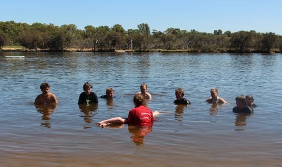 An instructor in the water with children gathered around as she demonstrates how to roll a victim over in the water