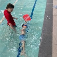 A swim instructor watching a young boy swim with a red kickboard