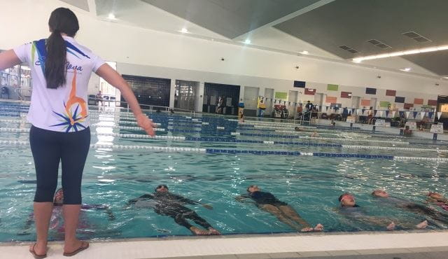 An synchro instructor by the side of the pool with a number of girls in the pool floating on their back