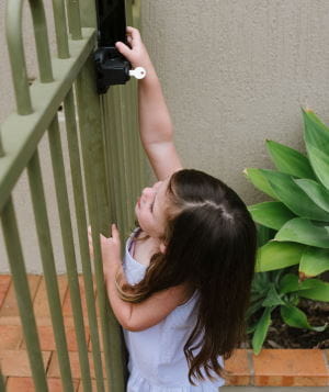 A toddler girl standing by a pool fence, reaching up to the latch.