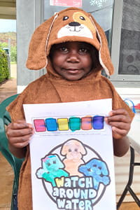 An Aboriginal child wearing a poncho towel and holding up their colouring page
