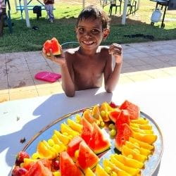 A young Aborginal boy holding fruit while standing beside a plate of fruit