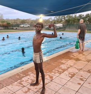 An Aboriginal boy standing by the pool with muscles flexed while other kids swim in the background