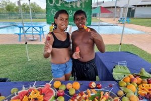 Two Aboriginal children standing at a table with fruit, eating fruit