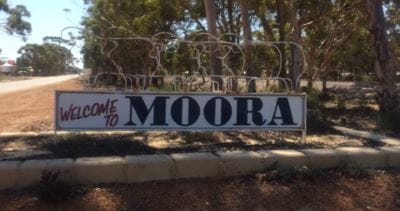 A sign with the town name Moora on it and trees along the roadside in the background
