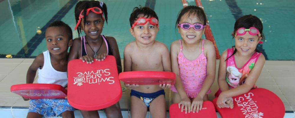 image of 5 multicultural children wearing swimming gear and sittin along the edge of a pool holding Swim and Survive kickboards