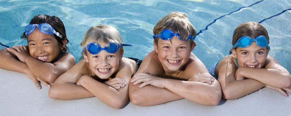 Four smiling children wearing blue goggles lean on the edge of a swimming pool