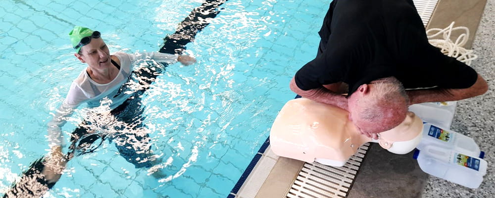 A woman in the pool while a man practises CPR on a manikin poolside