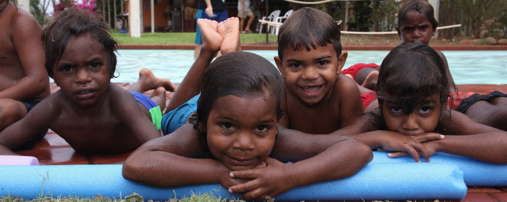 Aboriginal children laying on grass with pool noodles beside a swimming pool