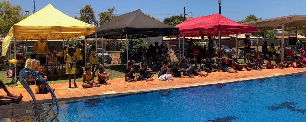 Children sitting under their yellow, black and red faction tents by  the pool at Bidyadanga