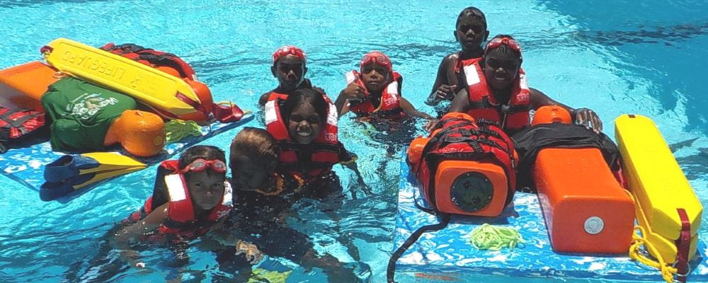 Seven aboriginal children in the pool at Bidyadanga wearing lifejackets and surrounded by rescue tubes and lifesaving manikins