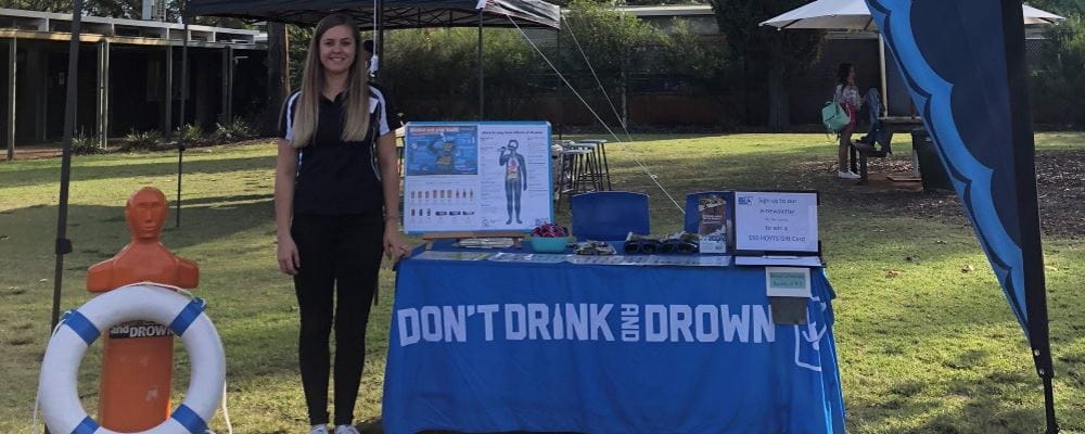 Don't Drink and Drown Co-ordinator Katie Cowcher standing by a presentation stand with Don't Drink and Drown merchandise and education materials on a school oval