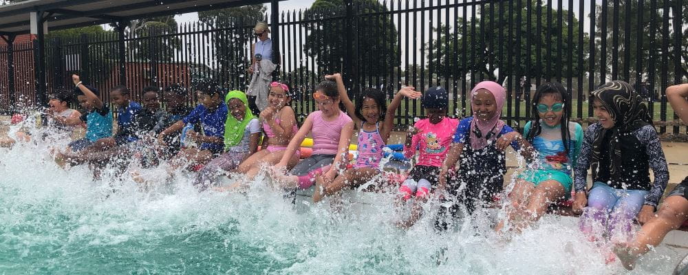 A group of multicultural children siting along the edge of a pool splashing their legs in the water