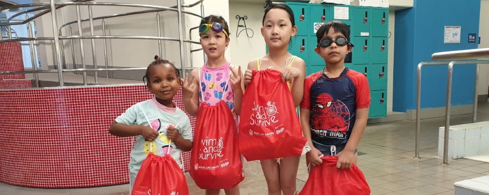 Four multicultural children holding red Swim and Survive bags on the pool deck at Leisurepark Balga