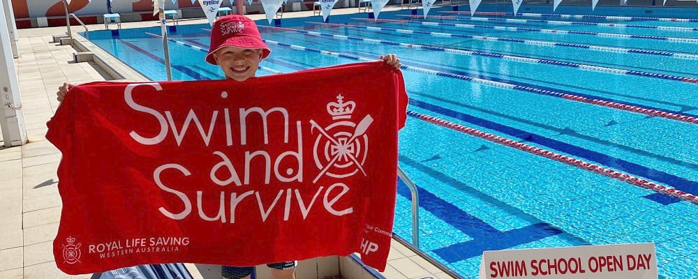 young boy standing in front of Churchlands pool holding Swim and Survive banner