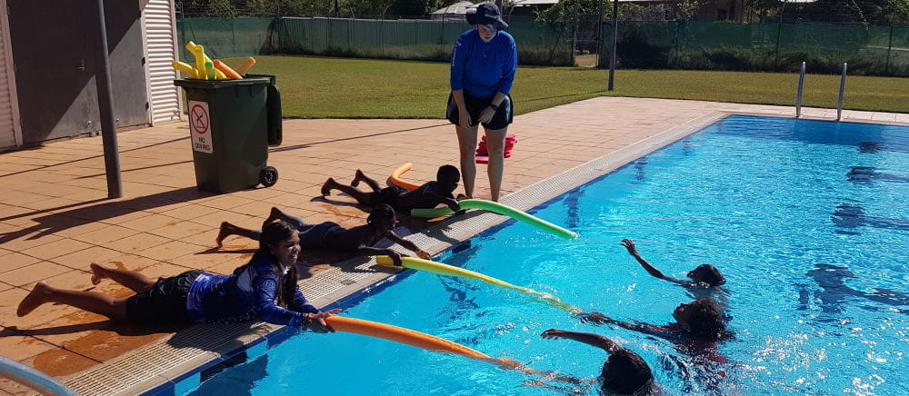 A swim instructor with a group of aboriginal girls using pool noodles to rescue their friends in the pool